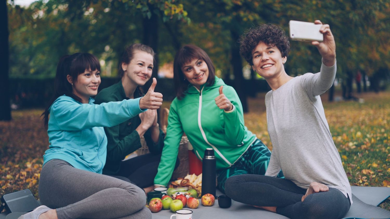 Four coworkers smiling and giving thumbs up on a picnic blanket in a park